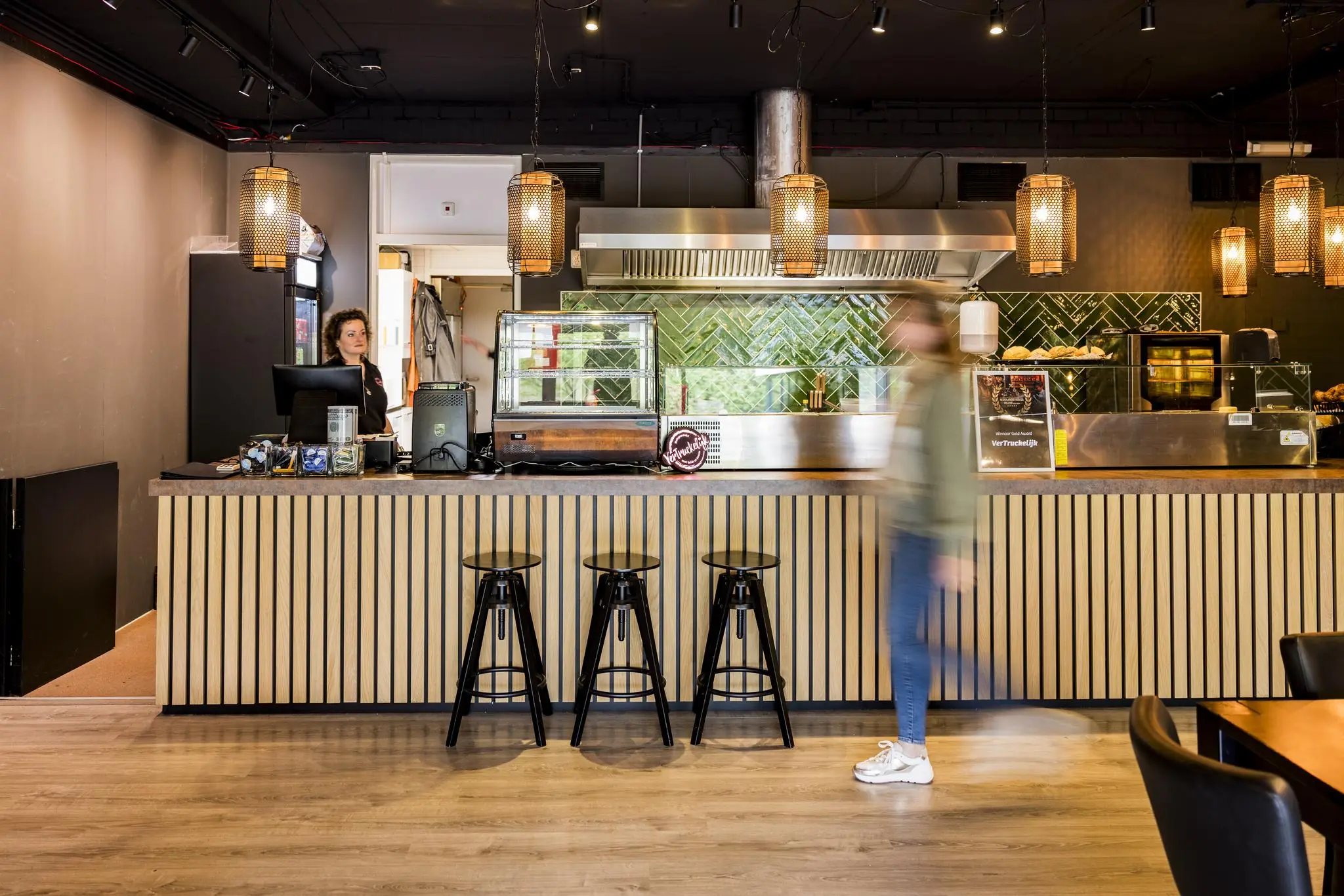 Modern ingerichte horecagelegenheid aan de Markerkant met houten bar, hanglampen en een medewerker achter de kassa.